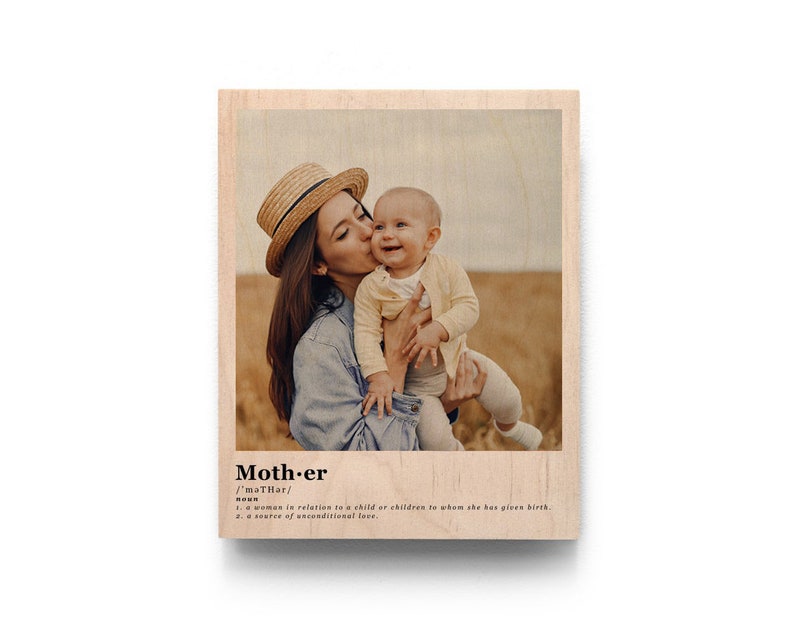 May include: A photo of a mother and child in a field. The mother is wearing a straw hat and a blue denim jacket. The child is wearing a white shirt and is being held by the mother. The photo is printed on a wooden plaque with the word "Mother" and its definition printed below the photo.