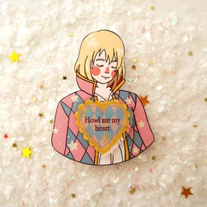 Può includere: Uno spillo illustrato rosa e blu di un personaggio con i capelli biondi e uno spillo a forma di cuore che dice "Howl ate my heart".