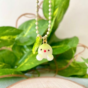 May include: A light green glass charm necklace with a small, round, light green charm with a red nose and two small white arms. The charm is attached to a light green chain.
