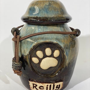 May include: A ceramic pet urn with a brown leather strap and a paw print design. The urn is glazed in shades of blue, brown, and white. The name "Reilly" is written on the front of the urn.