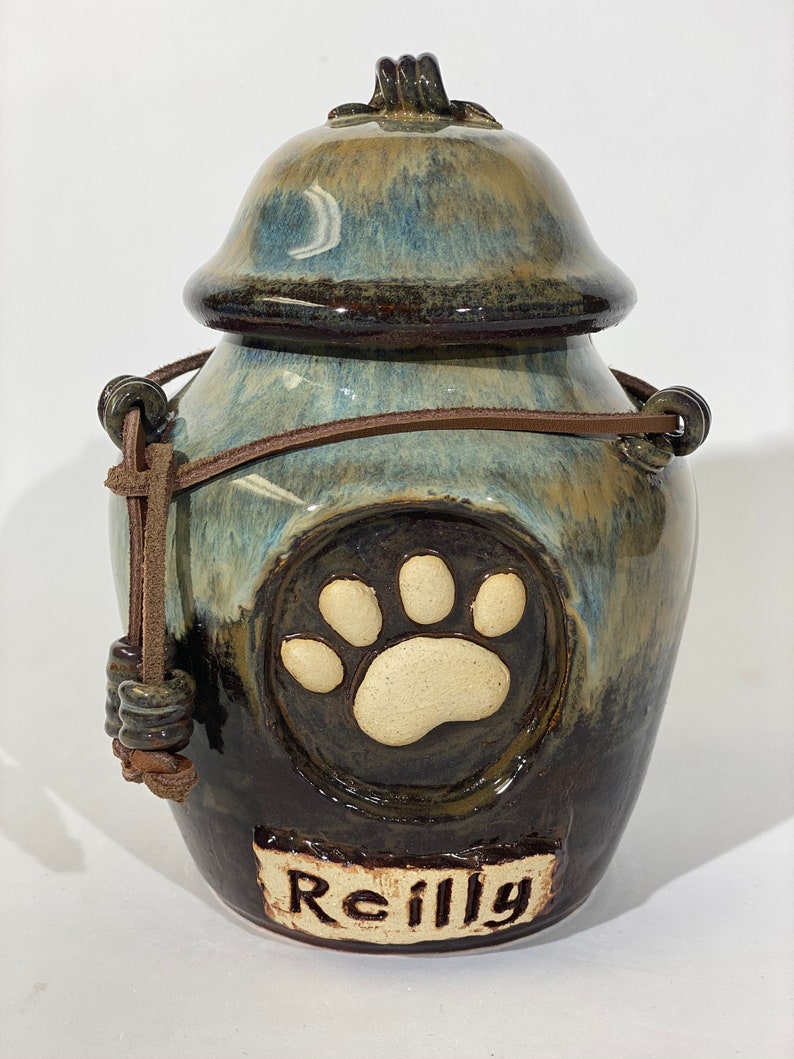 May include: A ceramic pet urn with a brown leather strap and a paw print design. The urn is glazed in shades of blue, brown, and white. The name "Reilly" is written on the front of the urn.