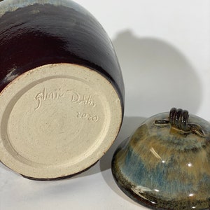 May include: A brown and blue glazed ceramic jar with a lid. The jar has a brown glaze with a blue and white glaze on the top. The lid has a brown glaze with a blue and green glaze on the top. The jar is signed "Janice Dotts 8220".