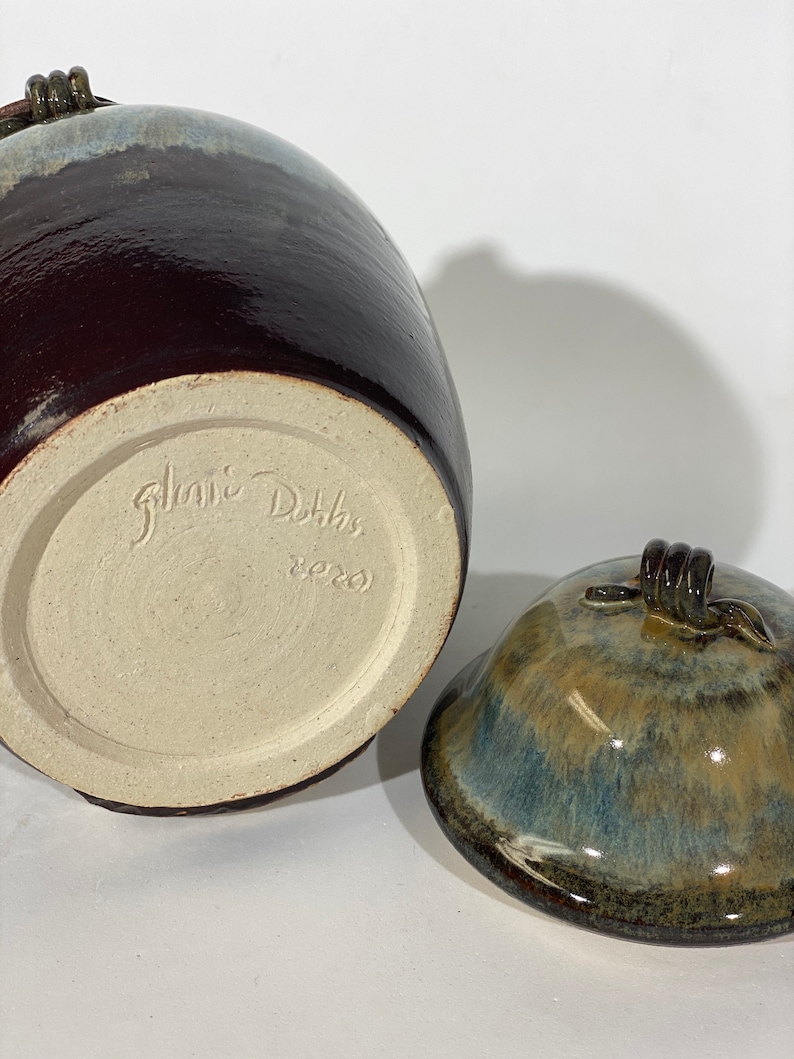 May include: A brown and blue glazed ceramic jar with a lid. The jar has a brown glaze with a blue and white glaze on the top. The lid has a brown glaze with a blue and green glaze on the top. The jar is signed "Janice Dotts 8220".