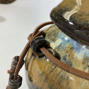 May include: A brown ceramic jar with a lid and a brown leather strap with dark brown beads. The jar has a mottled brown, blue, and black glaze.