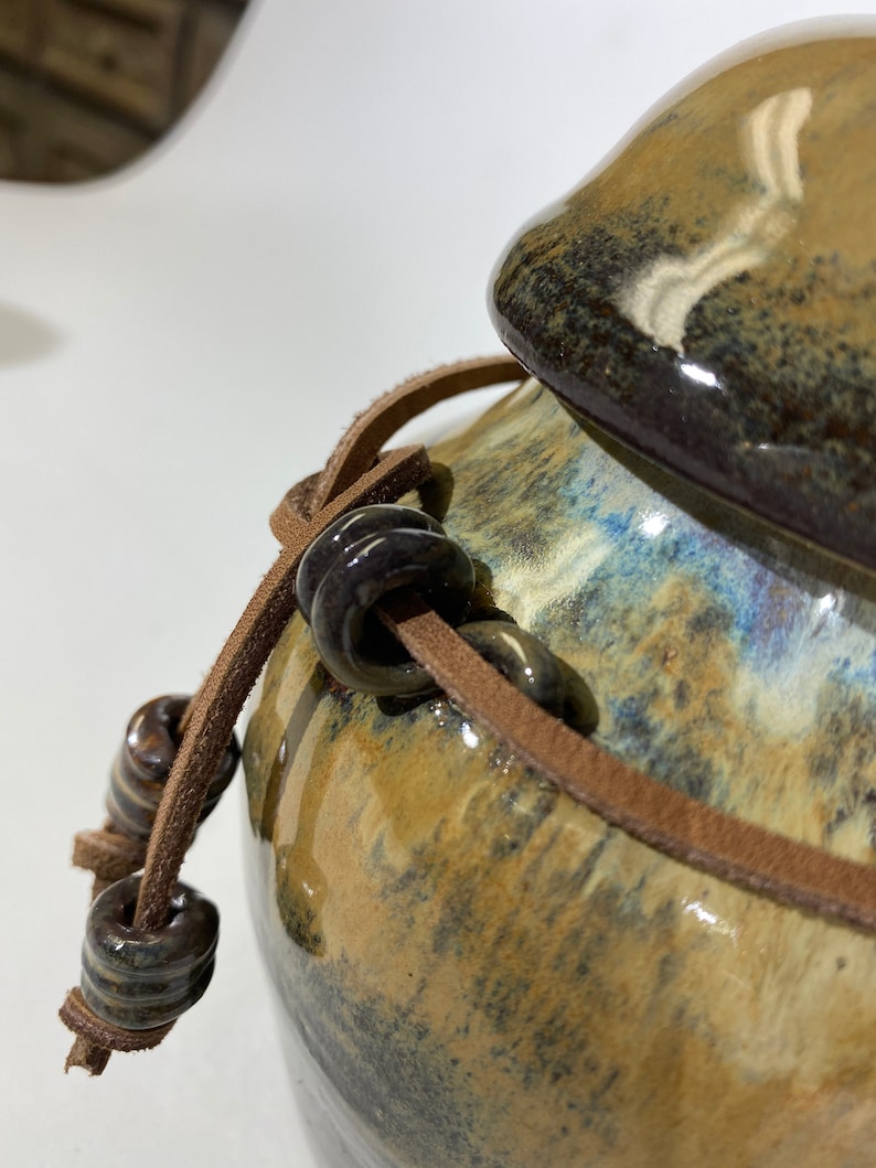 May include: A brown ceramic jar with a lid and a brown leather strap with dark brown beads. The jar has a mottled brown, blue, and black glaze.