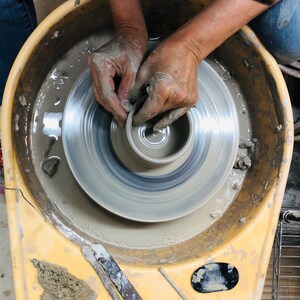 May include: A person's hands shaping a clay pot on a pottery wheel. The wheel is spinning and the clay is being molded into a bowl shape. The pottery wheel is in a yellow basin.