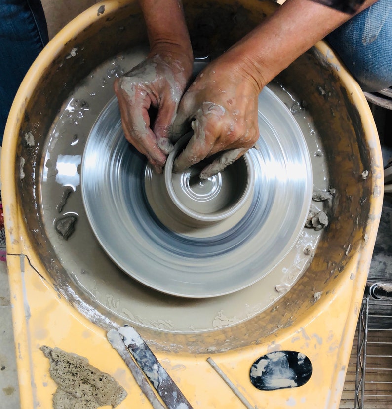 May include: A person's hands shaping a clay pot on a pottery wheel. The wheel is spinning and the clay is being molded into a bowl shape. The pottery wheel is in a yellow basin.