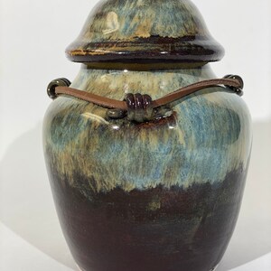 May include: A ceramic jar with a lid and a leather strap handle. The jar has a blue, brown, and white glaze.
