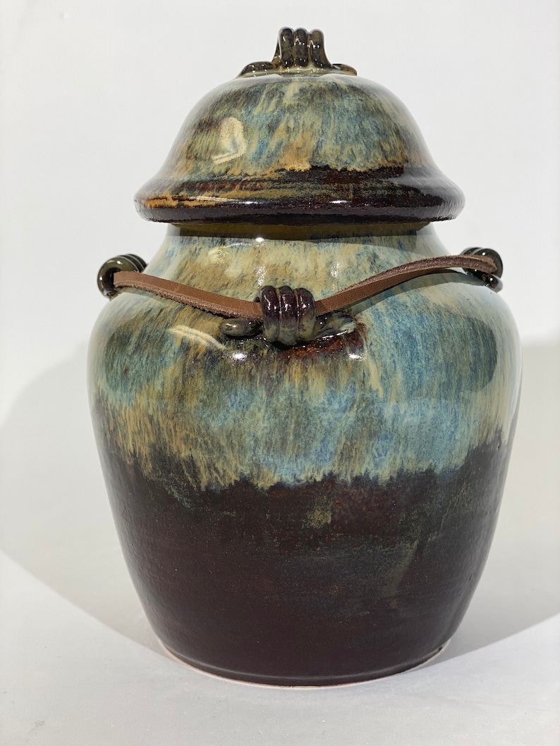 May include: A ceramic jar with a lid and a leather strap handle. The jar has a blue, brown, and white glaze.