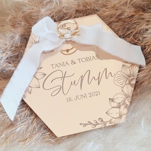 May include: A hexagonal wooden ring box with a floral design and the text "Tania & Tobias" and "Sturm" with the date "18. Juni 2021".