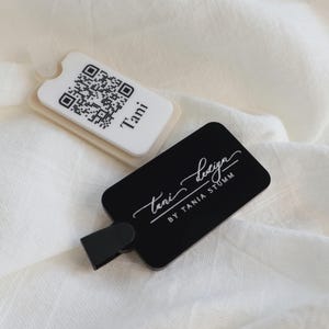 Könnte beinhalten: Zwei rechteckige Anhänger mit abgerundeten Ecken. Einer ist weiß mit einem QR-Code und dem Namen "Tani". Der andere ist schwarz mit dem Text "tani design BY TANIA STUMM" in weißer Schrift. An dem schwarzen Anhänger ist ein schwarzer Clip befestigt.