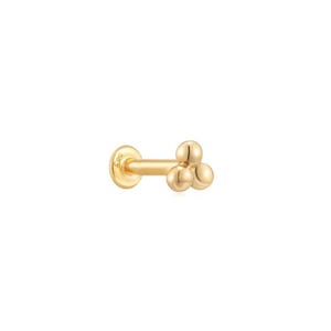 Pode incluir: Um brinco dourado com uma parte traseira plana e redonda e um pino reto. A frente do brinco apresenta três pequenas esferas redondas em relevo. O brinco é projetado para um piercing e provavelmente é feito de ouro.