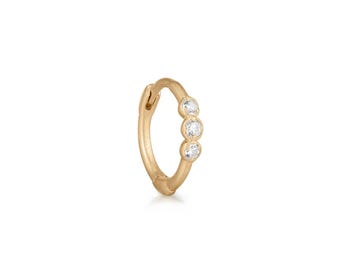 May include: A gold hoop earring featuring three small, clear, round-cut stones set in a vertical row along the outer edge. The hoop has a polished metallic finish and a hinged closure mechanism visible at the top of the circular band structure.