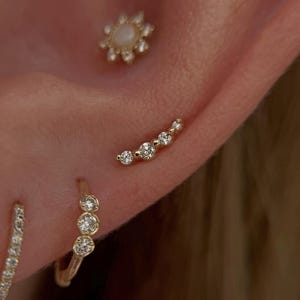 May include: A gold ear piercing set featuring a flower-shaped stud with a central opal and clear stones, a curved bar stud with five clear stones, and a small gold hoop earring adorned with three clear stones along the front edge.