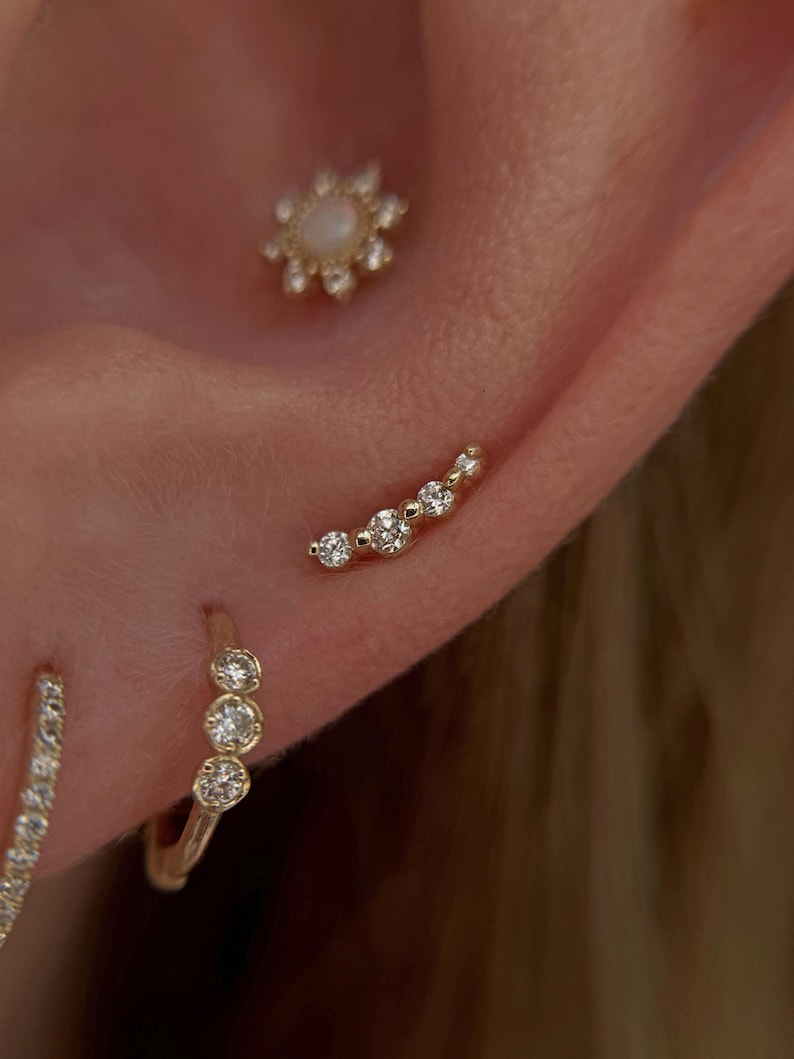 May include: A gold ear piercing set featuring a flower-shaped stud with a central opal and clear stones, a curved bar stud with five clear stones, and a small gold hoop earring adorned with three clear stones along the front edge.