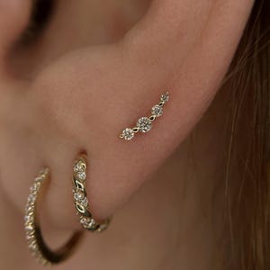 May include: A gold ear climber earring featuring five round clear stones arranged in a curved line, accompanied by two gold hoop earrings. One hoop has a twisted texture with small stones, and the other is a plain gold hoop with stones.