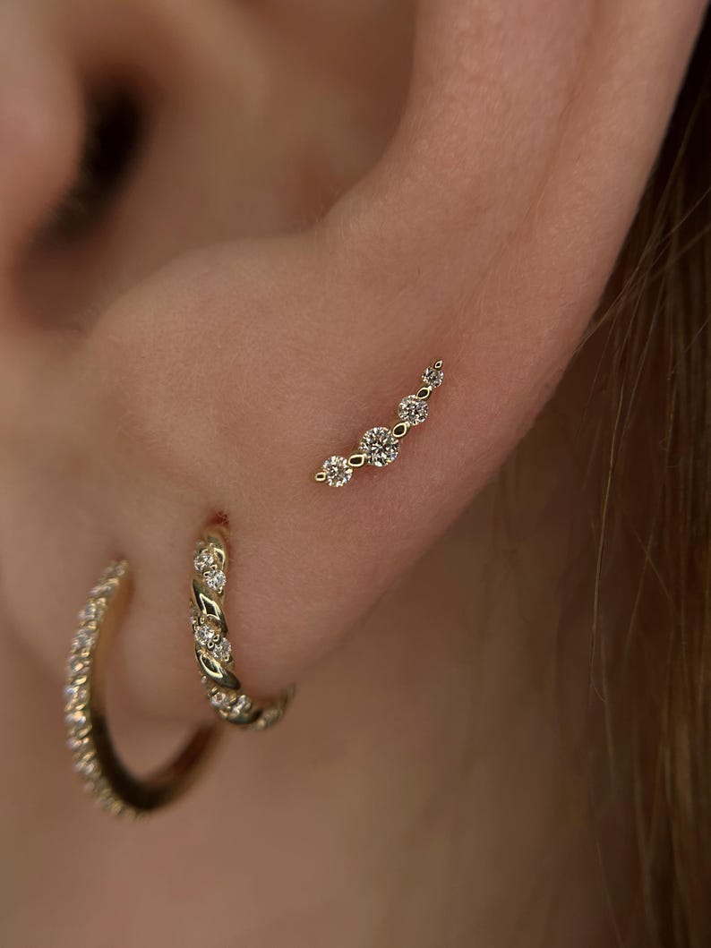 May include: A gold ear climber earring featuring five round clear stones arranged in a curved line, accompanied by two gold hoop earrings. One hoop has a twisted texture with small stones, and the other is a plain gold hoop with stones.