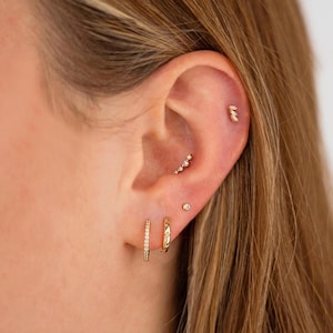 May include: A collection of gold-toned jewelry worn in the ear, featuring two small hoop earrings in the lobe, a single stud, a curved bar with multiple small stones in the conch, and a small triple-stone stud located in the upper cartilage area.