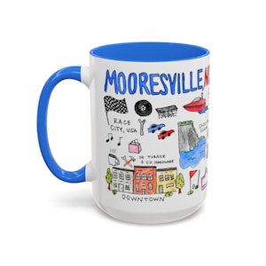 Peut inclure: Mug en céramique blanche avec intérieur et anse bleus. L'illustration colorée représente Mooresville, USA, avec le texte "MOORESVILLE" et des dessins de monuments et commerces locaux. La tasse est de taille standard.
