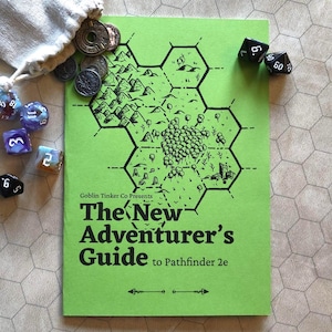 Könnte beinhalten: Ein grünes Heft mit dem Titel "The New Adventurer's Guide to Pathfinder 2e" mit einer Kartenillustration. Würfel, Münzen und ein kleiner Beutel sind um das Heft herum verstreut, was auf ein Tabletop-Gaming-Thema hindeutet.