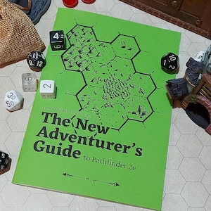 May include: A green booklet with the title "The New Adventurer's Guide to Pathfinder 2e" and a black and white illustration of a hexagonal map. The booklet is surrounded by dice.