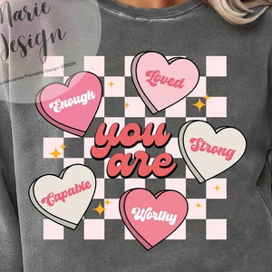 May include: A gray sweatshirt with a checkered pattern and pink hearts that say "You are" with the words "Loved", "Enough", "Strong", "Capable", and "Worthy" inside the hearts.
