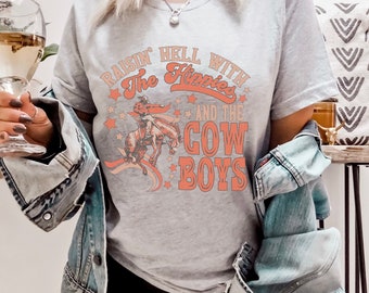 Könnte beinhalten: Ein graues T-Shirt mit dem Text "Raisin' Hell With The Hippies And The Cowboys" in Orange und Weiß. Das Shirt zeigt eine Vintage-Illustration eines Cowboys, der ein Pferd reitet.