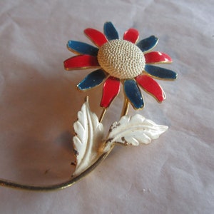 May include: A gold-toned brooch in the shape of a flower with red, blue, and white enamel. The flower has a white center with a textured surface and is attached to a gold stem with two white leaves.