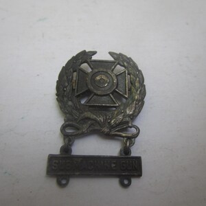 May include: A silver submachine gun expert badge with a wreath and the words "Submachine Gun" on a bar.