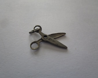 May include: A silver-toned metal charm in the shape of a pair of scissors. The charm has a loop at the top for attaching to a necklace or bracelet.