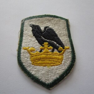 May include: A white embroidered patch with a green border. The patch features a black eagle perched on a gold crown.