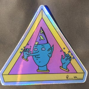May include: A holographic sticker featuring a blue character with a pointed hat and a long nose. The character is inside a pink triangle with a yellow border. The text "@___blh___" is at the bottom of the sticker.