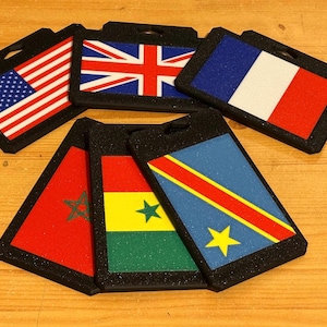 Puede incluir: Varios porta-credenciales negros, cada uno con una bandera nacional diferente. Se muestran las banderas de Estados Unidos, Reino Unido, Francia, Marruecos, Ghana y la República Democrática del Congo. Dispuestos sobre una superficie de madera.