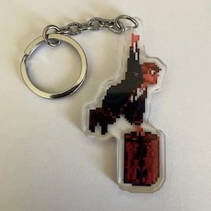 May include: A clear acrylic keychain featuring a pixel art design of a dark-haired figure in a black suit holding a red suitcase. The figure is depicted in a dynamic pose with one arm raised. The keychain includes a silver metal ring and chain.
