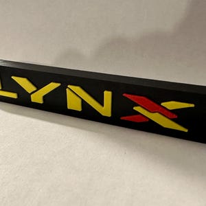 Può includere: Una placca di plastica nera con la parola "LYNX" in lettere gialle e rosse. Le lettere sono in rilievo e hanno un effetto 3D.