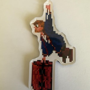 May include: A clear acrylic pin featuring a pixel art character with brown hair, wearing a dark blue jacket and holding a red flag. The character is positioned above a large, rectangular, dark red textured trunk with black hardware details.