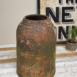 May include: A large, terracotta-coloured planter with a rough, textured surface. The planter has a green patina and is slightly weathered. It is sitting on a whitewashed wooden surface.