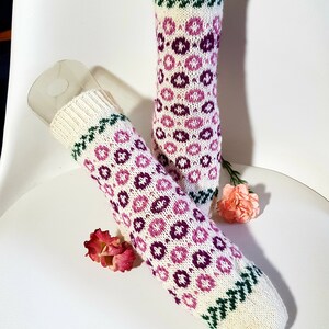 May include: A pair of white knitted socks with a pink and purple floral pattern. The socks have a green trim at the top and bottom.