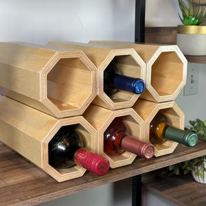 May include: A wooden wine rack with six octagonal compartments, each holding a wine bottle. The rack is made of light-colored wood and is displayed on a shelf. The bottles have various colored capsules.