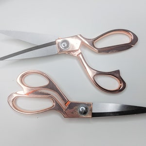 May include: Two pairs of rose gold scissors with silver blades are shown on a white background. One pair is open, the other closed. The scissors have a sleek design, suitable for crafting and sewing. The blades are approximately 10 cm long.