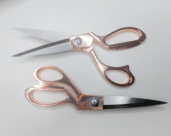 Könnte beinhalten: Zwei Paare roségoldener Scheren mit silbernen Klingen auf weißem Hintergrund. Ein Paar ist geöffnet, das andere geschlossen. Die Scheren haben ein elegantes Design, geeignet für Bastel- und Nähprojekte. Die Klingen sind ca. 10 cm lang.