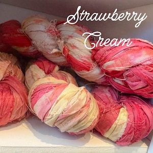 May include: Close-up of several skeins of fabric ribbon in shades of pink, red, and cream. The text "Strawberry Cream" is written in a cursive font across the top of the image. The ribbons appear to be made of a soft, textured material.