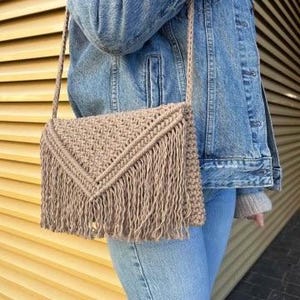 May include: A beige macrame crossbody bag with a fringed flap closure. The bag has a woven pattern and a long, braided strap. The bag is approximately 10 inches wide and 8 inches tall.