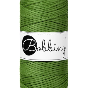 May include: A spool of green macrame cord with a white label that reads "Bobbiny" in a cursive font. The cord is tightly wound, showing a textured surface. Ideal for crafting projects.