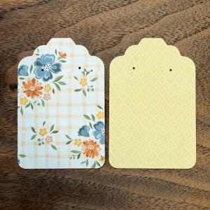 Pode incluir: Dois cartões retangulares para exibição de brincos com topos recortados e dois pequenos orifícios. O cartão esquerdo apresenta um padrão xadrez bege claro com motivos florais azuis, laranja e amarelos. O cartão direito tem um padrão de treliça amarelo.