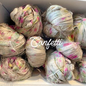May include: A collection of bundled fabric ribbons in various sizes. The ribbons are primarily beige with pink and yellow floral patterns. The word "Confetti" is written in a cursive font across the image.