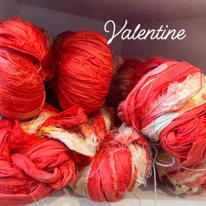 May include: A collection of red fabric bundles, some tightly wound and others loose, with a mix of textures. The word "Valentine" is written in a cursive font across the top. The fabric appears to be silk or a similar material.