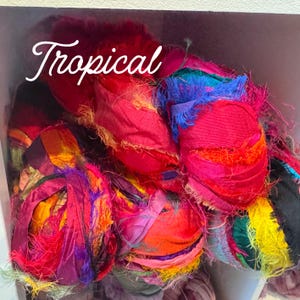 May include: A collection of colorful fabric bundles in shades of red, pink, orange, yellow, and blue. The word "Tropical" is written in white script across the top of the image. The fabric appears to be textured and fringed.