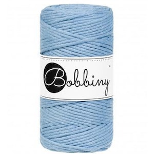 May include: A cylindrical spool of light blue macrame cord. The cord is tightly wound and wrapped with a white label featuring the brand name "Bobbiny" in a black, cursive font. The cord is suitable for various crafting projects.