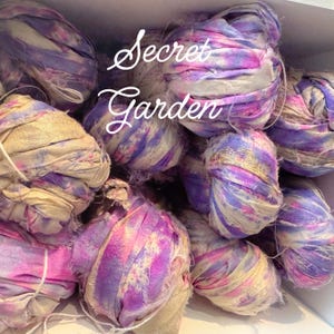 May include: A collection of colorful fabric ribbon bundles in shades of purple, pink, and cream. The text "Secret Garden" is written in a cursive font across the top of the image. The ribbons appear to be made of silk or a similar material.